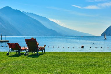 Plajda şezlong üzerinde oturan ve yaz saati, görünümü dağlarla çevrili güzel Lake Garda Garda Gölü hayran insanlar