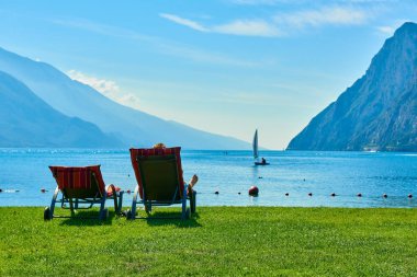Plajda şezlong üzerinde oturan ve yaz saati, görünümü dağlarla çevrili güzel Lake Garda Garda Gölü hayran insanlar