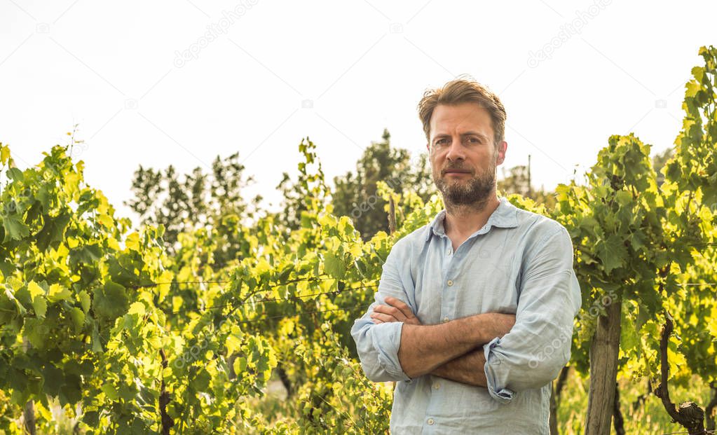 Serious forty years old caucasian farmer standing proud in front of a vineyard. Agriculture or gardening - country outdoor scenery, warm sunset light.