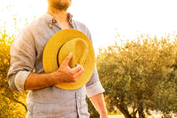 オリーブ畑の前に立ちながら麦わら帽子をかぶった白人農家や庭師 農業やガーデニング 国の屋外風景 金の夕日の光 — ストック写真