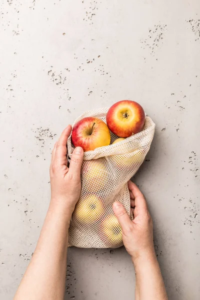 Hands holding apples in a eco friendly (zero waste, plastic free) cotton mesh bag. Captured from above (top view, flat lay). Grey concrete background - copy space.