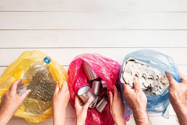 Recycling and ecology. People sorting (segregating) household waste (paper, metal, plastic, glass) into bags captured from above (flat lay, top view). White wooden background.