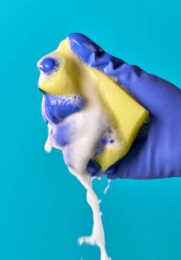 Eldivenli elini sarı hijyenik sünger sıkıştırma. Su, köpük. Mavi arka plan.