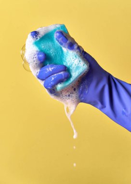 Eldivenli elini mavi hijyenik sünger sıkıştırma. Su, köpük. Sarı arka plan.