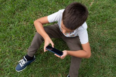Küçük çocuk çimlerin üzerinde yatay pozisyonda cep telefonu kullanarak oturuyor. Tam üstten..