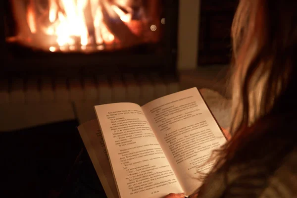 young girl reads a book in front of a fireplace in the evening at home