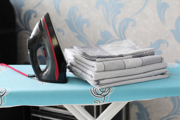 A black iron stands on the ironing board. Near a pile of bed linen.