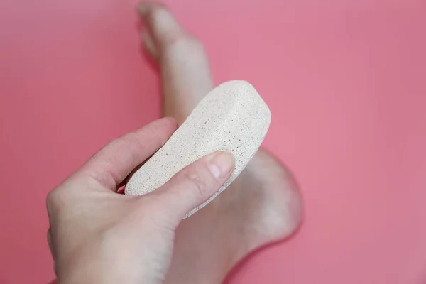 In the photo a woman hand holds a pumice near the heel of her foot.