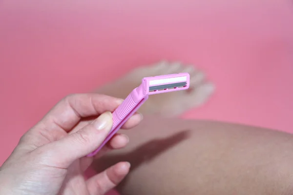 Beautiful female hand holding a pink razor against the background of the legs. — Stock Photo, Image