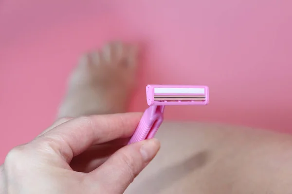 Beautiful female hand holding a pink razor against the background of the legs. — Stock Photo, Image
