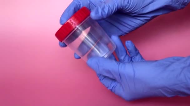 一只戴蓝色胶乳手套的手拿着一个收集尿液的容器 可供医生分析的塑料容器 分析器是空的 粉色背景 — 图库视频影像