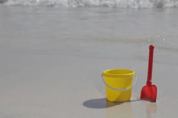 Un cubo de bebé amarillo y una pala roja están en la arena junto al mar . — Foto de Stock