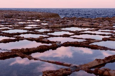 Gozo, Malta 'da buharlaşma metoduyla deniz tuzu üretim noktası. Doğal elmas şekilli havuzlar, gün batımında dalgaların bıraktığı suyla. Pembe gökyüzü ve gölde yansıyan bulutlar