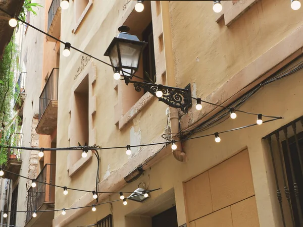 Lighting lamps on wire hangs outside against cozy facade as a festive decoration. Narrow beautiful street of Barcelona, Spain