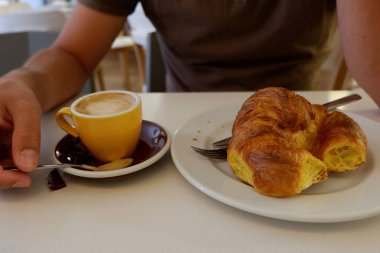 Bir insan bir kafedeki küçük bir masada oturur, sıcak kruvasanın tadını çıkarır ve zengin bir fincan kahveyi yudumlar. Atmosfer rahat ve davetkar. Sabah yemeği için mükemmel..