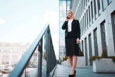 Ciddi bir iş kadını ofis binasının yanında dikilirken akıllı telefondan konuşuyor.