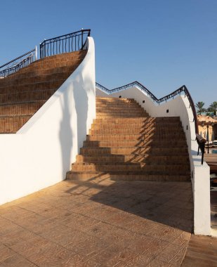 beyaz bir duvar ve mavi berrak gökyüzüne karşı metal dekor ile tuğla adımlar ve ahşap korkuluklar ile iki katmanlı merdiven