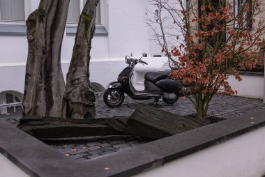 Scooter, sokakta beyaz bir duvarın arka planda ağaçlar arasında moped