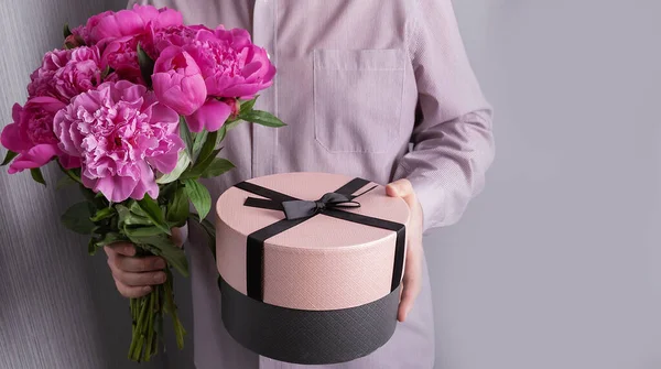 Yuvarlak kutulu bir adam hediye ve şakayıklı bir buket tutuyor. Pembe çiçekler ve gri arka planda hediye.