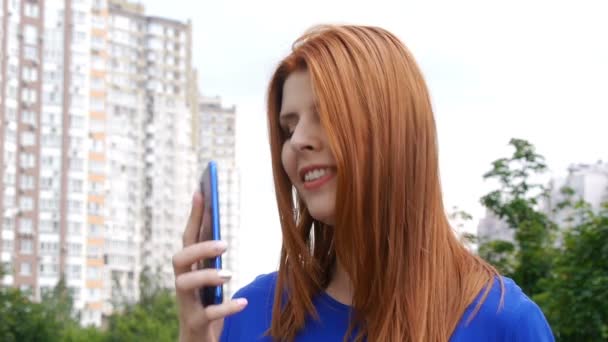 市内に赤い髪をした若い美しい女の子は、電話番号をダイヤルし、携帯電話で話して笑顔. — ストック動画
