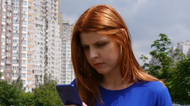 Menina bonita nova com cabelo vermelho, na cidade, não consegue encontrar informações em um telefone celular . — Vídeo de Stock