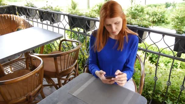 Sehr schönes Mädchen mit roten Haaren sitzt in gemütlichen Café. mit dem Telefon, lächelnd. — Stockvideo