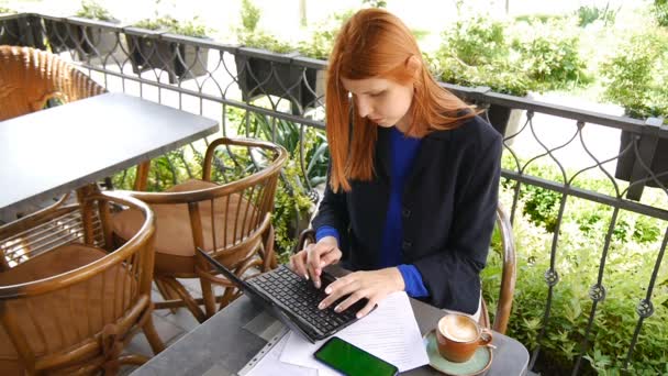 Jovem mulher de negócios atraente usando laptop no café. Documentos, telefone celular com tela verde e xícara de café na mesa. Mulher de cabelo vermelho em uma jaqueta . — Vídeo de Stock