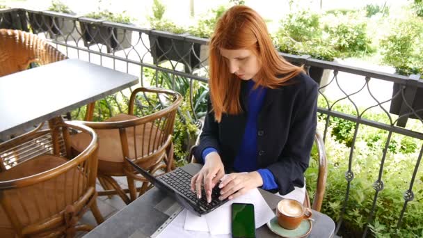 Junge attraktive Geschäftsfrau mit Laptop und Handy mit grünem Bildschirm im Café. Dokumente und eine Tasse Kaffee auf dem Tisch. Frau mit roten Haaren und Jacke. — Stockvideo