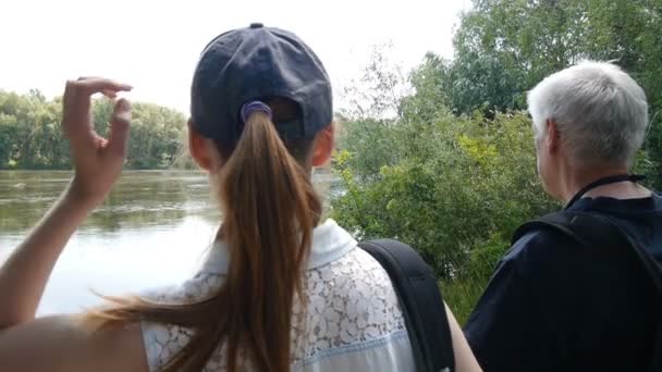 El hombre y la niña caminan juntos. Senderistas con mochilas. Se detienen en la orilla del río y lo miran. Chica tocando y corrigiendo su pelo largo . — Vídeos de Stock