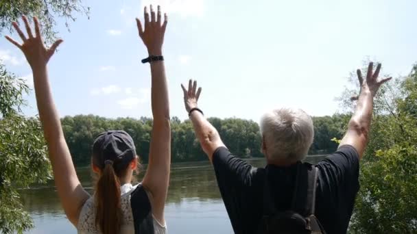 Concepto de viajes felices. El hombre y la niña caminan juntos. Los excursionistas con mochilas se detienen en la orilla del río y lo miran. Son felices y levantan las manos. . — Vídeos de Stock