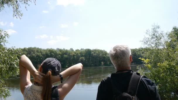 Concepto de viajes felices. El hombre y la niña caminan juntos. Los excursionistas con mochilas paran en la orilla del río y se relajan. Son felices y levantan las manos. . — Vídeo de stock