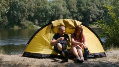 İçerik mutlu çift çadırda oturan. Turizm ve seyahat kavramı. Kız termostan çay döküyor. Arka planda orman ve nehir.