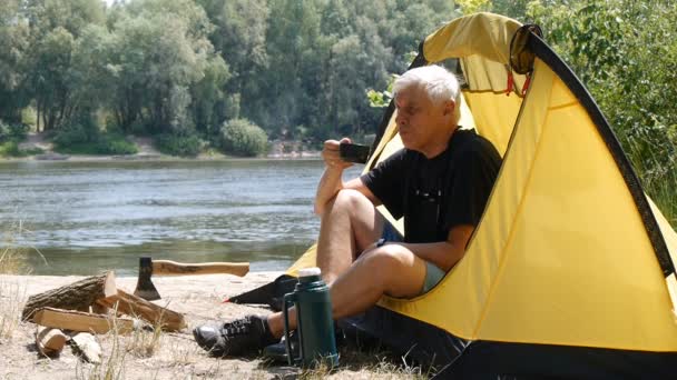 Hiker sittande i tält. Camper man dricka kaffe eller te. Floden och skogen i bakgrunden. Avslappning, Resor, grön turism koncept. — Stockvideo