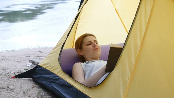 Молодая женщина отдыхает и читает книгу в палатке на природе. Река на заднем плане. Пешие прогулки, путешествия, зеленый туризм концепции. Здоровый активный образ жизни . — стоковое видео