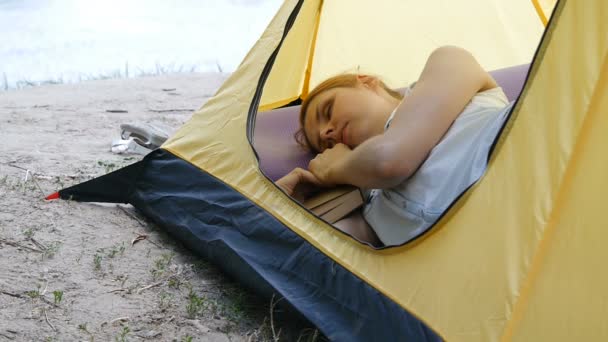 Una joven viajera duerme en una tienda. Chica senderista cansado se quedó dormido con un libro en una tienda de campaña. Senderismo, viajes, concepto de turismo verde. Vida activa saludable . — Vídeo de stock