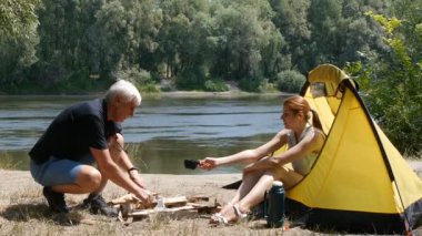 Genç kadın gezgin çadırda oturuyor. Kız adama bir fincan çay geçirir. Adam çay içiyor ve gülüyor. Yürüyüş, seyahat, yeşil Turizm konsepti. Arka planda nehir ve orman.
