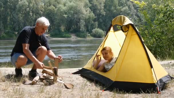 Fiatal nő utazó alszik a sátorban. Egy férfi közeledik a sátor és elindul aprító fa a tüzet. A lány felébred. Túrázás, utazás, zöld turizmus koncepciója. A háttérben a folyó és az erdő. — Stock videók