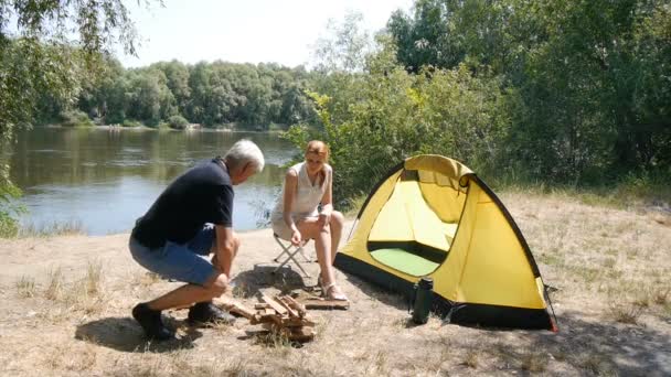 Un hombre y una chica están sentados junto al fuego. La chica lanza leña al fuego. Turismo, viajes, concepto de turismo verde. Río y bosque en el fondo . — Vídeo de stock