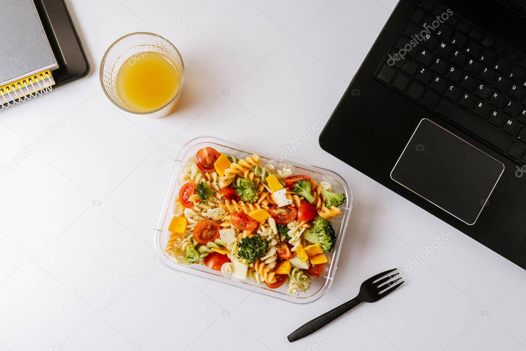 Healthy food in lunch box, on working table with laptop .Ensalada de pasta brocoli con queso y verduras. eating at workplace. Home food for office concept 