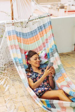 Yaz günü, gülümseyen bir kadın çatıda renkli bir hamakta, cep telefonu kullanıyor. Kadın blogu, sohbet ya da internetten alışveriş, dijital aletler. Yaşam tarzı..
