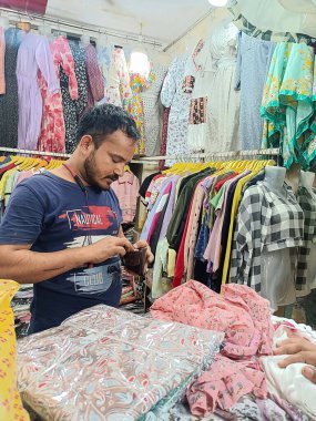 Kolhapur, Hindistan 3 Eylül 2023 Mavi tişörtlü bir adam tezgahın arkasında dururken küçük bir cüzdanı ve parayı kontrol ediyor. Renkli elbiselerle çevrili, küçük, parlak bir giyim mağazasında..