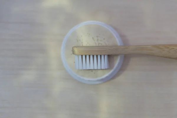 Natural bamboo toothbrush with wooden handle and white bristles lies on a round box with natural toothpaste and a light wooden background under natural light with sun glare top view — Stock Photo, Image