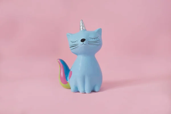 Juguete de recuerdo de cerámica moneybox gatito Korn azul con cola de arco iris colorido con ojos cerrados y cuerno de unicornio sobre fondo rosa en luz natural Fotos de stock libres de derechos