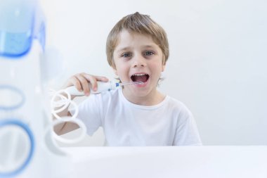 5 yaşındaki bir çocuk beyaz bir t-shirt beyaz bir masada evde oturur ve bir oral tahriş edici ile dişlerini yıkarsıyor. çocuk gülümsüyor ve sinirli kolu tutar.