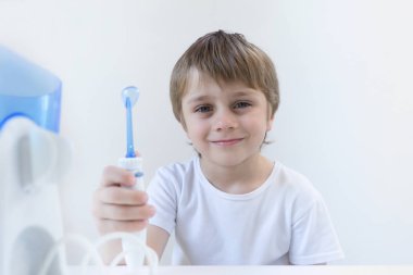 5 yaşındaki bir çocuk beyaz bir t-shirt beyaz bir masada evde oturur ve bir oral tahriş edici ile dişlerini yıkarsıyor. çocuk gülümsüyor ve sinirli kolu tutar.
