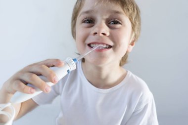 5 yaşındaki bir çocuk beyaz bir t-shirt beyaz bir masada evde oturur ve bir oral tahriş edici ile dişlerini yıkarsıyor. çocuk gülümsüyor ve sinirli kolu tutar.