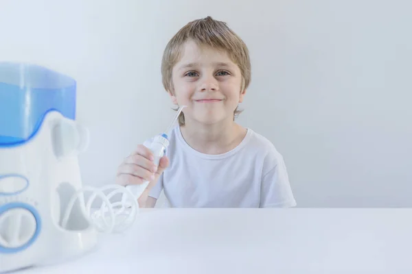 a 5-year-old boy sits at home at a white table in a white t-shirt and washes his teeth with an oral irrigator. the boy smiles and holds the handle of the irrigator.