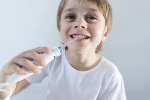 Seorang anak laki-laki berusia 5 tahun duduk di rumah di meja putih dalam t-shirt putih dan mencuci giginya dengan irrigator oral. anak itu tersenyum dan memegang pegangan dari iririgator . — Stok Foto