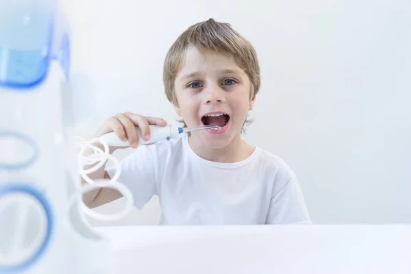 Un niño de 5 años se sienta en casa en una mesa blanca con una camiseta blanca y se lava los dientes con un irrigador oral. el niño sonríe y sostiene el mango del irrigador . Imágenes de stock libres de derechos