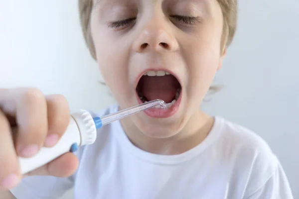 Un niño de 5 años se sienta en casa en una mesa blanca con una camiseta blanca y se lava los dientes con un irrigador oral. el niño sonríe y sostiene el mango del irrigador . Fotos de stock libres de derechos